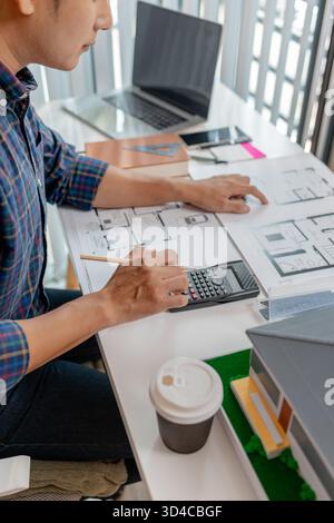 A young architect is working at his desk, reviewing blueprints, using a calculator, and checking a house model. He focuses on precision and detail whi Stock Photo