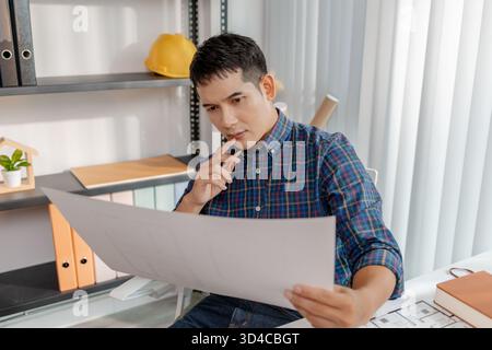 A young architect is working at his desk, reviewing blueprints, using a calculator, and checking a house model. He focuses on precision and detail whi Stock Photo