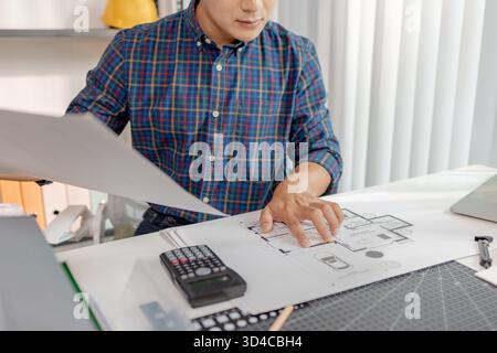 A young architect is working at his desk, reviewing blueprints, using a calculator, and checking a house model. He focuses on precision and detail whi Stock Photo