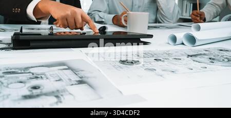A diverse business team engages in an architectural project, utilizing a digital tablet while reviewing detailed blueprints and designs in a modern Stock Photo