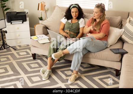 Discussing project Diverse female friends sitting on sofa in home office, with laptop and papers Stock Photo