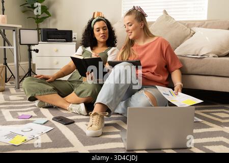 Diverse female friends studying documents in living room, with laptop, tablet and sticky notes Stock Photo