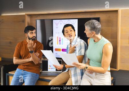 Discussing Diverse coworkers analyzing data on flat-screen at office desk, with printed documents Stock Photo