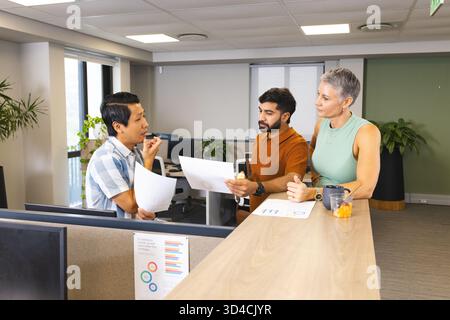 Collaborating Diverse coworkers reviewing printed charts at office desk, with coffee mug and plants Stock Photo