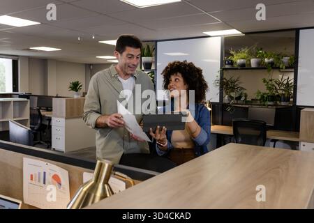 Diverse coworkers collaborating over printed papers and tablet at workstation, with potted plant Stock Photo