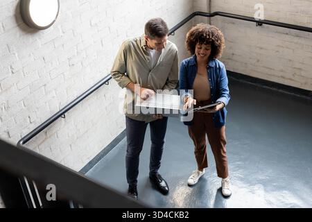 Discussing Diverse coworkers holding laptop and tablet on office stairs, by wall light and railings Stock Photo