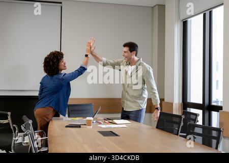 High-fiving Diverse coworkers celebrating teamwork in office conference room, with laptop notebook Stock Photo