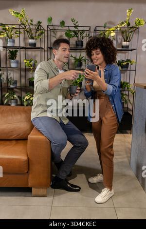 Diverse coworkers pointing at smartphone screen in office lounge, with mug and plant shelves Stock Photo