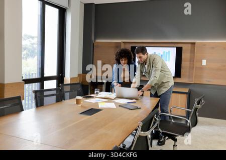 Reviewing printed charts, Diverse coworkers leaning over laptop in conference room, with coffee cup Stock Photo