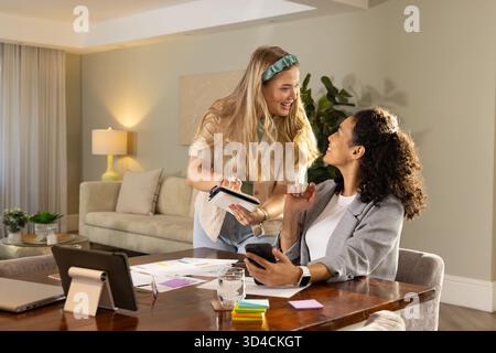 Collaborating female colleagues reviewing printed charts on table in home office, with smartphone Stock Photo