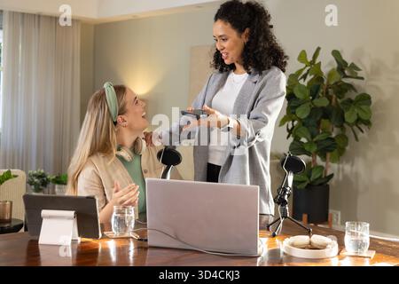 Collaborating Diverse female coworkers working at wooden table in home office, with laptop and mic Stock Photo