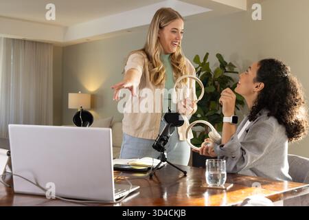 Passing headphones between Diverse female colleagues at home office desk with laptop and microphone Stock Photo