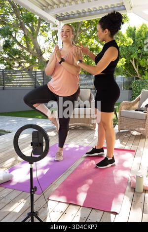 Practicing Diverse female trainer and client balancing on pergola deck, with yoga mats, ring light Stock Photo