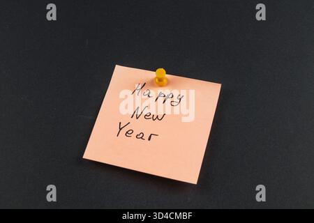 A simple sticky note with the message Happy New Year is pinned on a dark surface, symbolizing joy and new beginnings for the upcoming year. Stock Photo