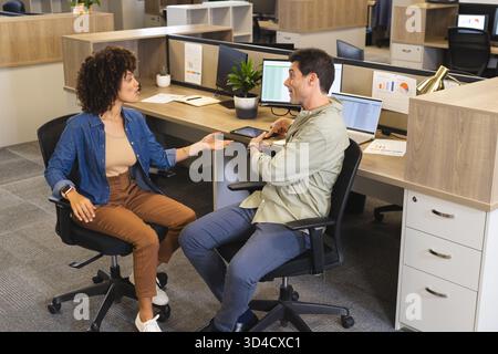 Discussing project charts, diverse coworkers leaning at office desk with two laptops tablet, plants Stock Photo