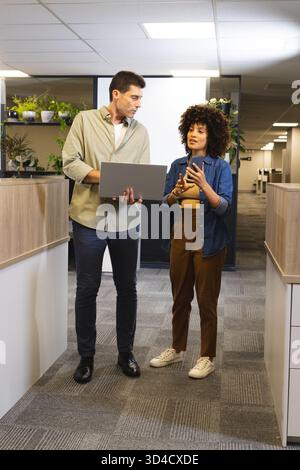 Talking Diverse coworkers standing in office corridor by plants, cubicles, with laptop smartphone Stock Photo