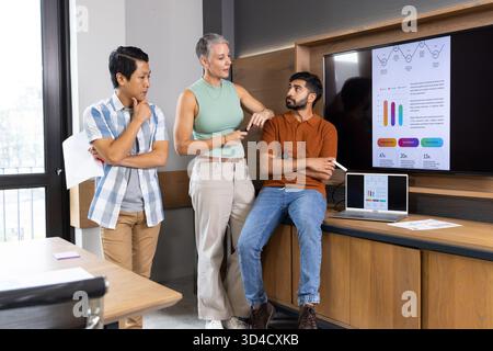 Discussing Diverse colleagues reviewing data on countertop in breakout area, with laptop and screen Stock Photo