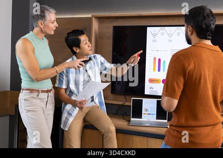 Collaborating diverse coworkers analyzing charts on wall monitor in office, with printed documents Stock Photo