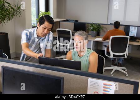 Collaborating Diverse coworkers reviewing printed chart on monitor at cubicles, with potted plants Stock Photo