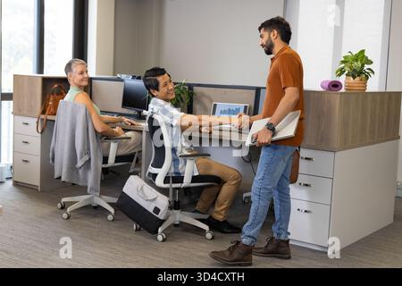 Indian and Asian male coworkers shaking hands across desk in open-plan office, with closed laptop Stock Photo