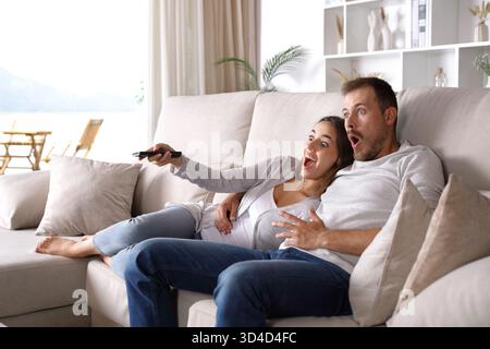 Amazed couple watching surprising tv program sitting on a couch at home Stock Photo