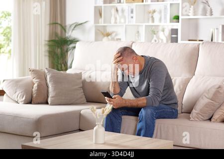 Worried senior man checking bad news on phone sitting on a couch at home Stock Photo