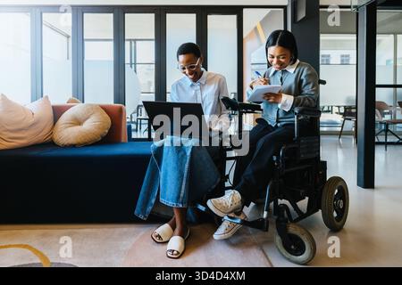 Two young women working together, one using a wheelchair, in a contemporary workspace Stock Photo