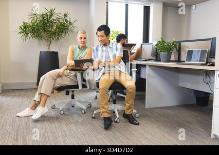 Collaborating Diverse coworkers reviewing content on rolling chairs in office, with digital tablet Stock Photo