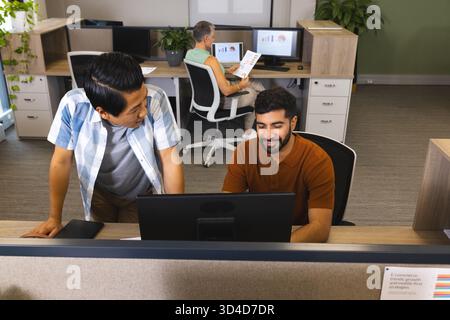 Collaborating diverse coworkers leaning over desk at office, with monitors and printed charts Stock Photo