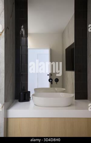 Interior of a modern and luxurious bathroom. It features a white ceramic vessel sink on a white countertop, placed on a wooden vanity. The walls are d Stock Photo