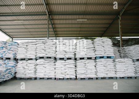 Neatly stacked white polypropylene bags arranged on wooden pallets in a covered warehouse. The industrial storage area features a metal roof structure Stock Photo