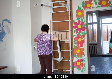 A woman in a purple floral top and brown pants adds a white attachment to stall bars fixed on a wooden board. The room has flower paintings on the wal Stock Photo