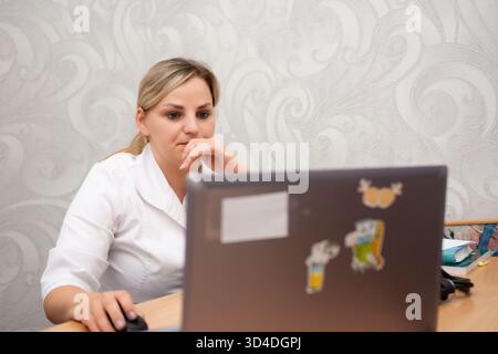 A fair-skinned woman, possibly a doctor, is focused on a laptop, wearing a white lab coat. She is in an office space with a decorative patterned wallp Stock Photo