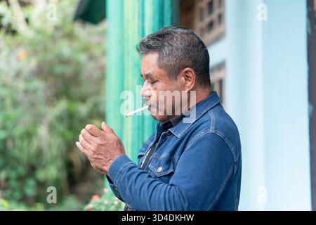 A candid portrait of a senior Asian man smiling while holding a cigarette. He stands outside his home with blue walls in a rural village in Indonesia. Stock Photo