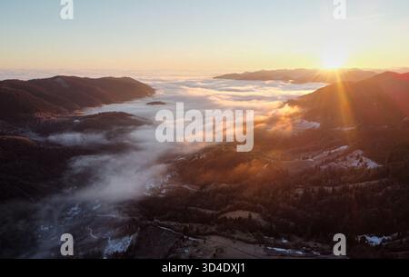 Setting sun casts golden hue over vast sea of clouds, blanketing valleys between forested hills and distant mountain ranges. Aerial view of tranquil scene captures serene beauty of nature. Stock Photo