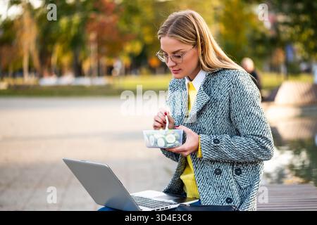 Modern businesswoman with eyeglasses in coat using laptop and enjoying  a healthy lunch break while sitting on a park bench in the city during sunny a Stock Photo