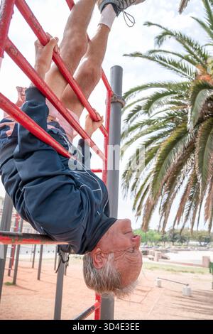 Senior man working out, hanging upside down on a red bar in an outdoor park Stock Photo