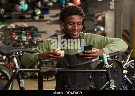 Young Black Man Eating Sandwich While Using Smartphone in Bike Workshop Stock Photo