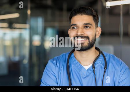 Young male doctor or medical professional with a beard smiling, looking away with a thoughtful or hopeful expression, wearing blue scrubs and a stetho Stock Photo