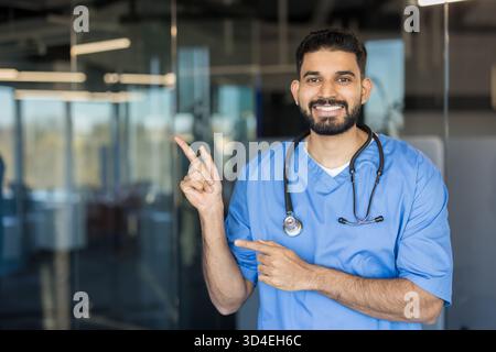 Young indian male doctor in blue scrubs and stethoscope smiling while using both hands pointing to the left, indicating good medical service and infor Stock Photo