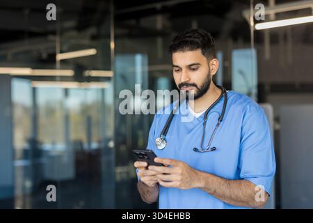 Young male doctor in scrubs and stethoscope typing on a smartphone, engaging in digital healthcare and medical communication within a hospital or clin Stock Photo