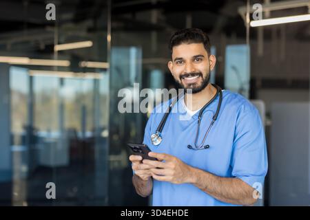 Smiling male doctor wearing blue scrubs and stethoscope, cheerfully looking at camera while holding and using a smartphone in a modern clinic or hospi Stock Photo
