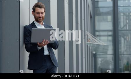 Successful mature Caucasian business man with laptop working in city leaning on wall of office company building businessman male employer developing Stock Photo