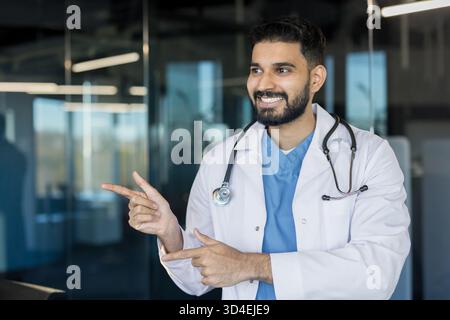 Young indian male doctor in a white coat with stethoscope, smiling confidently and pointing in a modern clinic office, suggesting professional guidanc Stock Photo