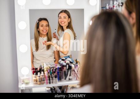 Beautician at work. Young woman is working as makeup artist in her beauty studio. She is applying eye shadow. Both women wear neutral-colored tops and Stock Photo