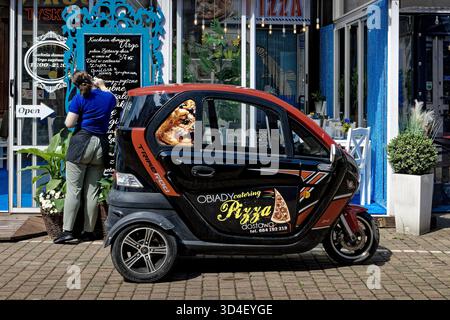 Pizza delivery microcar parked outside a restaurant, with a waitress writing the menu on the blackboard. Stock Photo