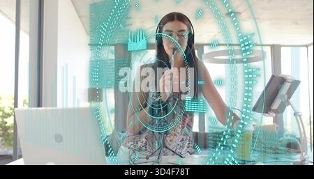 Speaking woman wearing sleeveless top and headphones, holding mic in bright office HUD treadmill Stock Photo