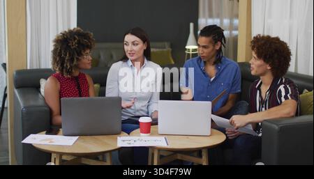 Leading woman in white blouse gesturing and guiding team on gray sofa with laptops and charts Stock Photo