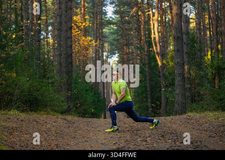Male runner in sportswear performing a lunge stretch on a forest path, focusing on warming up muscles and maintaining a healthy lifestyle while exercising in nature during autumn Stock Photo