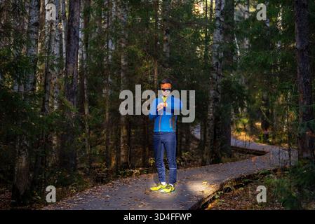 Man in athletic apparel is checking his smart watch after a training session on a wooden boardwalk trail surrounded by a dense forest, focusing on health and fitness data in nature Stock Photo